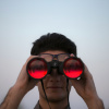 Young man peers through red-tinted binoculars held up to his eyes against a pale blue sky backdrop