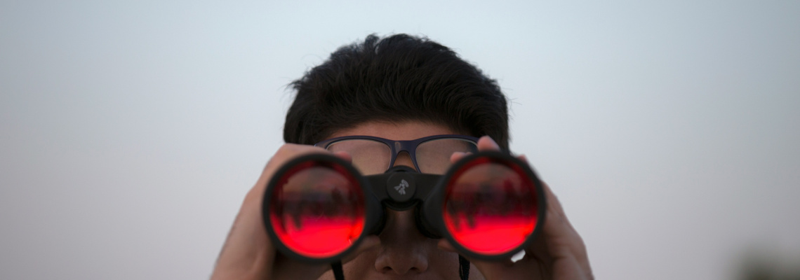 Young man peers through red-tinted binoculars held up to his eyes against a pale blue sky backdrop