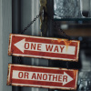 Rusty red 'ONE WAY' sign with a left-pointing white arrow hanging on chains between metal posts, blurred background behind.