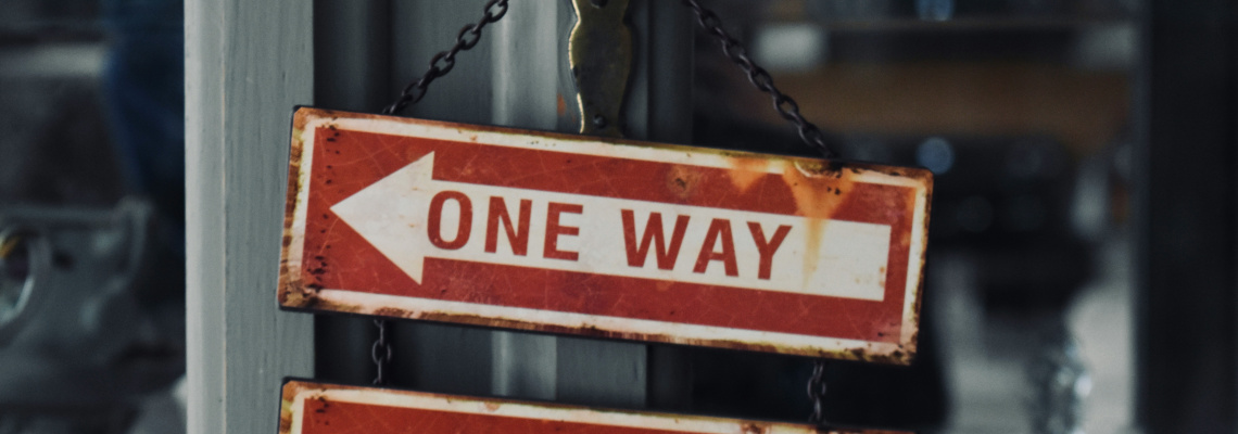 Rusty red 'ONE WAY' sign with a left-pointing white arrow hanging on chains between metal posts, blurred background behind.