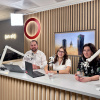 Three podcast hosts sit at a desk with microphones and laptops in a modern studio, wood-paneled backdrop and an “on air” sign behind them.
