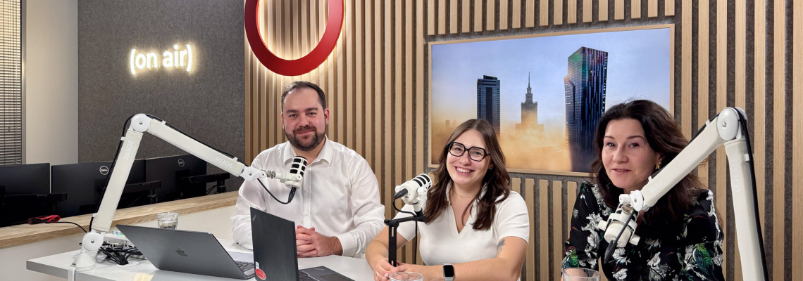 Three podcast hosts sit at a desk with microphones and laptops in a modern studio, wood-paneled backdrop and an “on air” sign behind them.