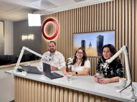 Three podcast hosts sit at a desk with microphones and laptops in a modern studio, wood-paneled backdrop and an “on air” sign behind them.